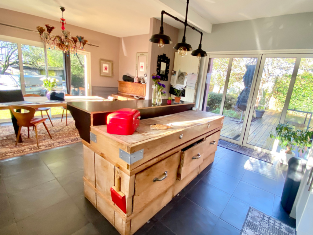 Une cuisine et un coin repas modernes dans cette maison à vendre à Saint Père en Retz comprennent un grand îlot en bois, des suspensions, un grille-pain rouge et une table à manger adjacente près de grandes portes coulissantes en verre menant à une terrasse extérieure.