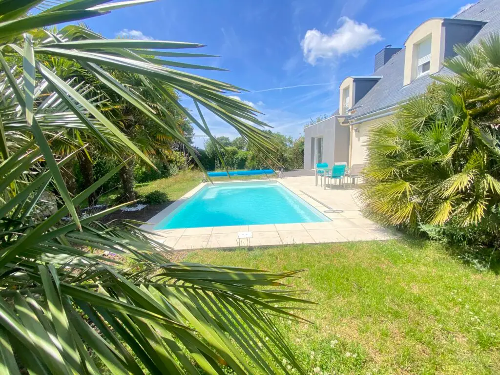 Une maison à vendre à Pornichet dispose d'une piscine dans la cour à côté d'une maison de couleur claire, entourée de palmiers et de verdure, sous un ciel bleu avec des nuages épars. Des carreaux de pierre blanche encadrent la piscine, créant un refuge idyllique.